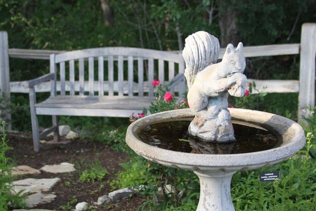 A small, stone fountain in a backyard garden, surrounded by lush plants and flowers. The fountain has multiple tiers and the water is gently cascading down, creating a relaxing atmosphere.