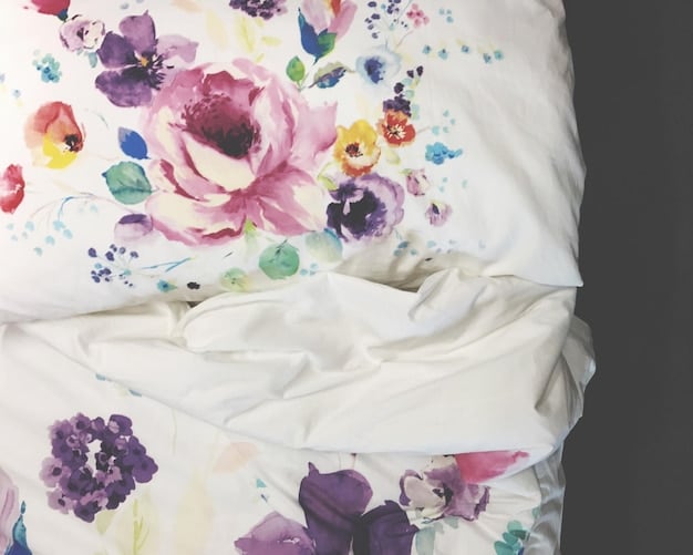 A close-up of a bed with patterned bedding. The duvet cover features a large-scale floral print in muted tones, while the throw pillows include a mix of smaller geometric and striped patterns. The overall effect is cozy and inviting.