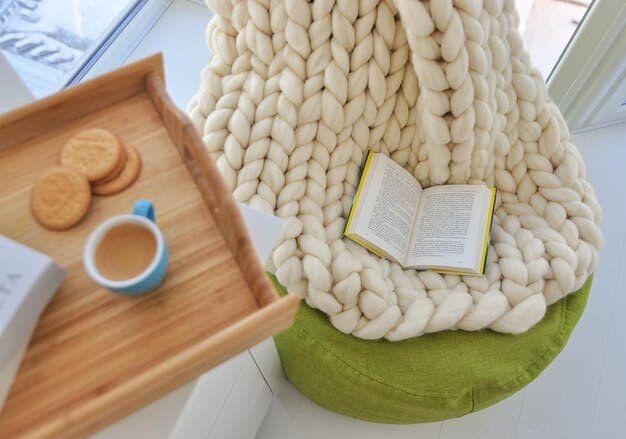 A close-up of a reading nook featuring a plush, oversized armchair with a knitted throw blanket, a small side table holding a cup of tea and a stack of books, and a warm, inviting lamp providing soft lighting.